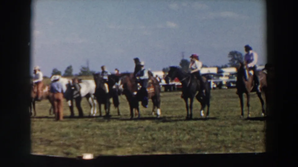 Wider 1957 Wheeling, Ohio frame with multiple riders and horses assembled on grass before or during a horse racing event.