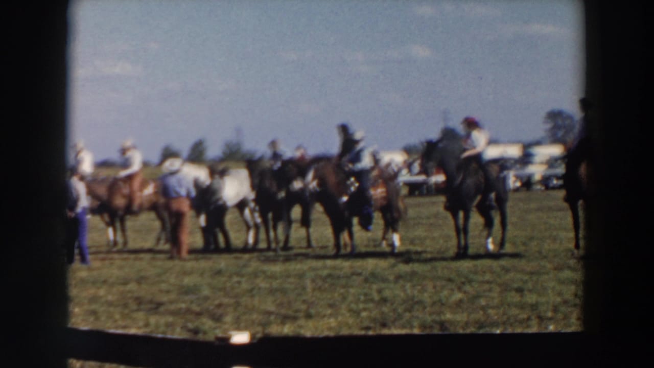 Riders on horseback gather in a grassy field with people standing nearby.