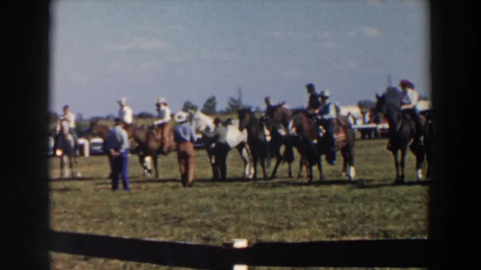 1957 Wheeling, Ohio rodeo footage frame with riders on brown, black, and light-colored horses, surrounded by standing attendees and distant parked cars.