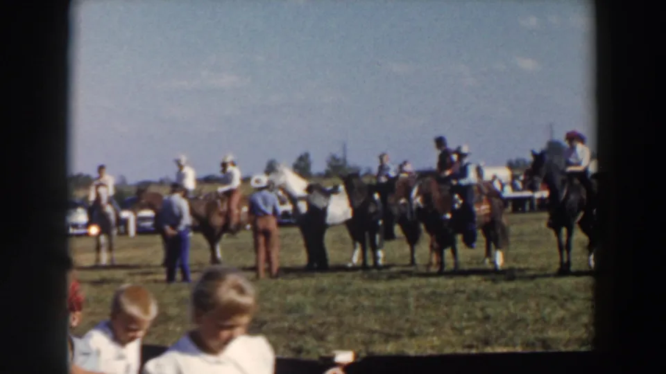 1957 Wheeling, Ohio 8mm rodeo frame showing riders and horses in the field, with two young spectators partly visible in the foreground and cars behind the activity.