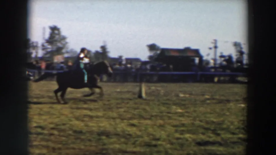 1957 Wheeling, Ohio rodeo frame showing a rider on a dark horse galloping or trotting across the grassy field, with a fence, spectators, vehicles, and buildings in the background.