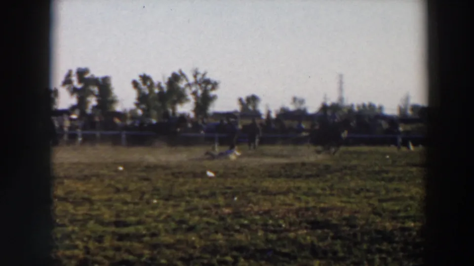 1957 Wheeling, Ohio home-movie frame showing a rodeo rider on horseback near lassoed livestock in a grassy arena with spectators beyond the fence.