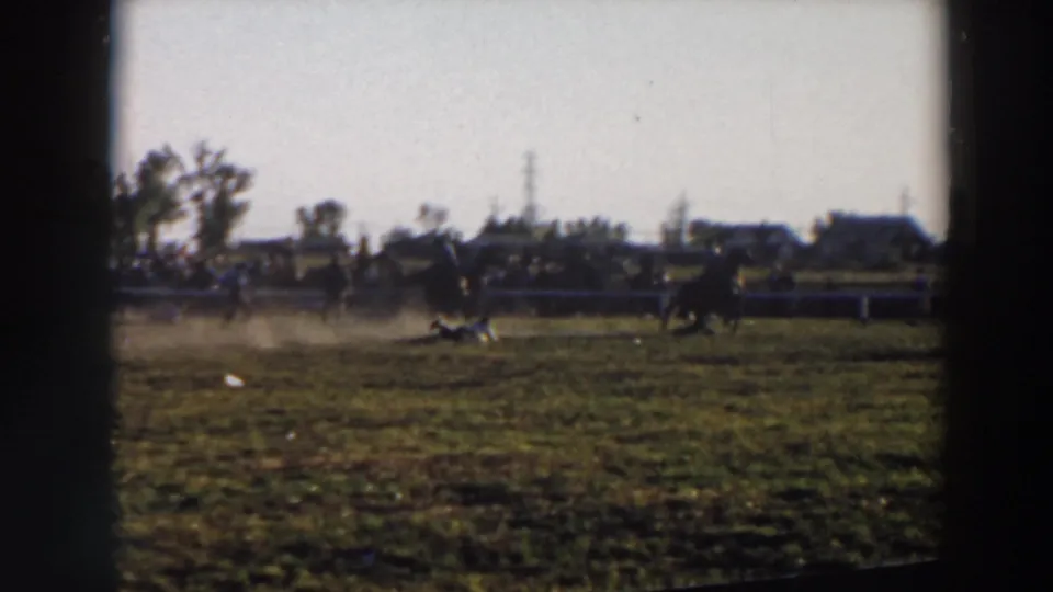 Wide 8mm frame from the Wheeling, Ohio rodeo showing the rider and livestock action across a fenced grassy field under a bright sky.