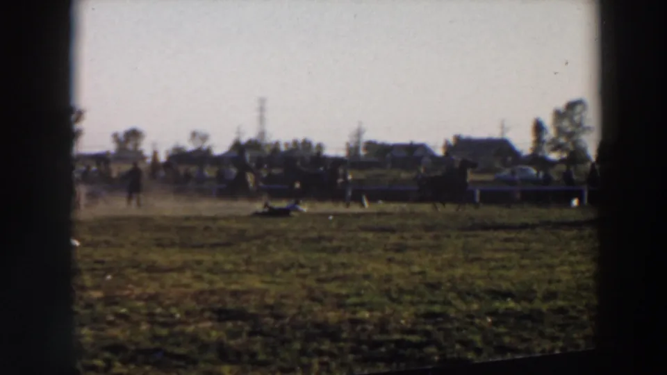 1957 Wheeling, Ohio rodeo frame showing dusty livestock handling in a grassy arena with distant spectators, trees, and houses beyond the fence.