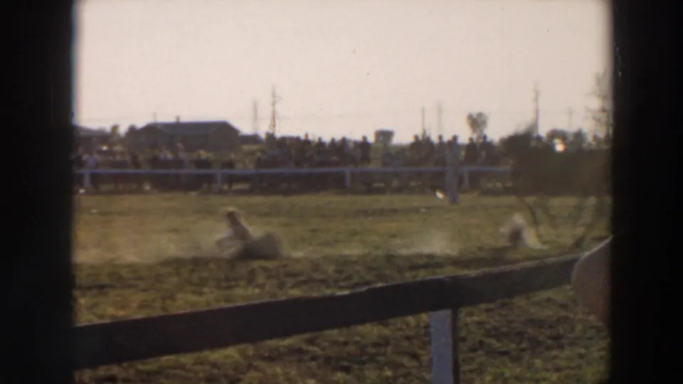 1957 Wheeling, Ohio home-movie frame showing a person down on the grassy rodeo field with spectators lined behind the fence in bright daylight.