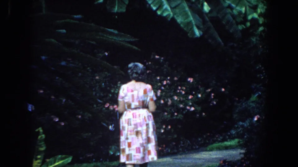 In supplied 1958 Florida home-movie footage, a woman in a pink-and-white print dress walks away along a lush garden path.