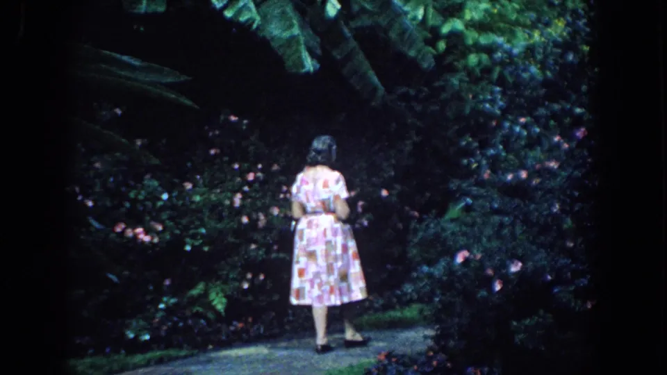 The woman pauses on the Florida garden path, looking toward flowering bushes amid dense greenery in grainy 8mm home-movie color.