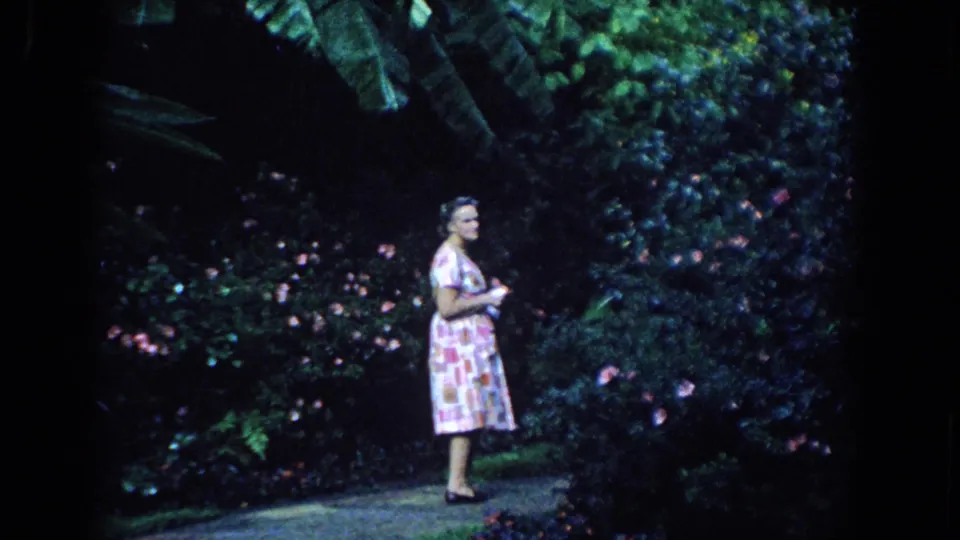 Near the end of the 1958 Florida garden shot, the woman turns partly toward the camera while standing among flowering shrubs and tropical greenery.