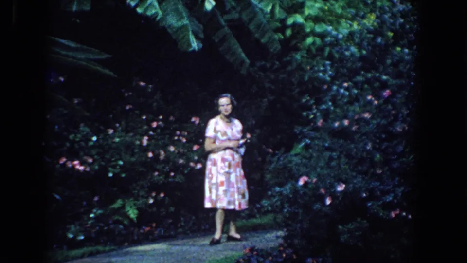 In 1958 Florida 8mm footage, a woman in a pink patterned dress stands on a shaded garden path near dense flowering plants.