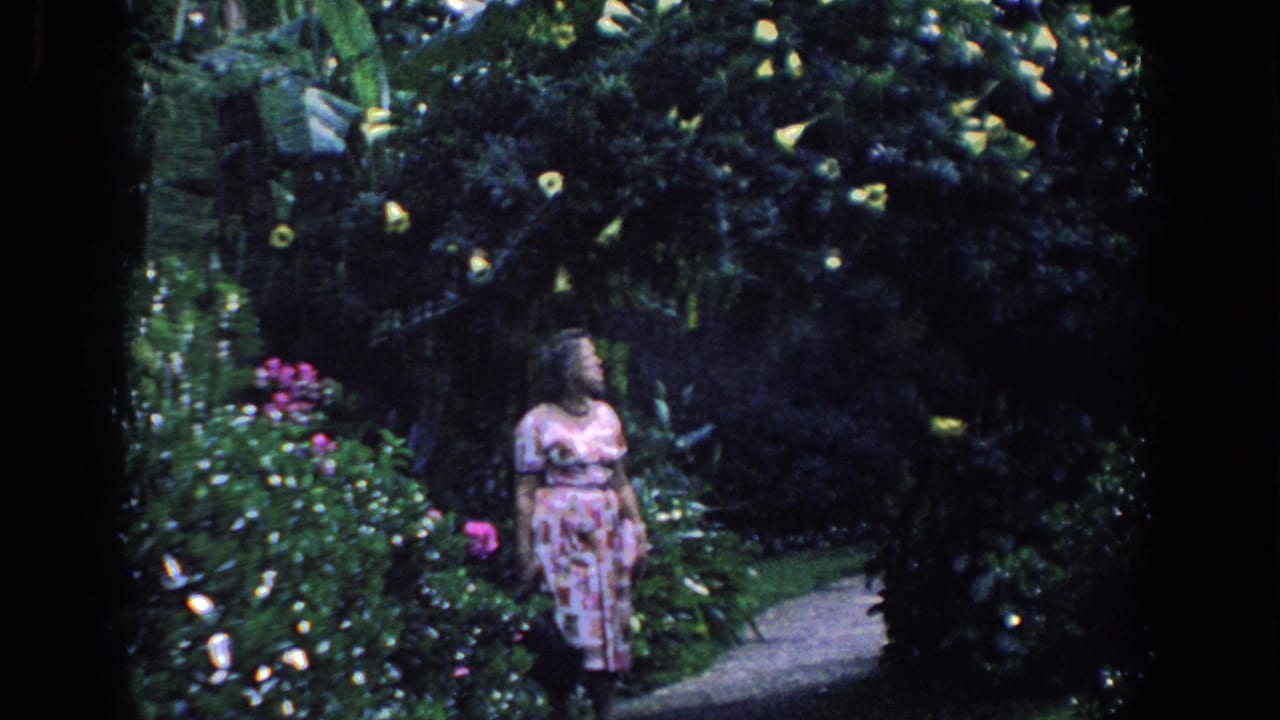 A woman in a pink patterned dress stands in a lush Florida garden with flowers and tropical leaves.