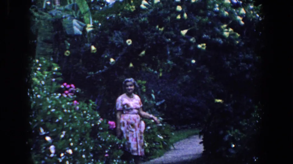 A woman in a pink patterned dress pauses on a Florida garden path surrounded by tropical foliage and flowers.