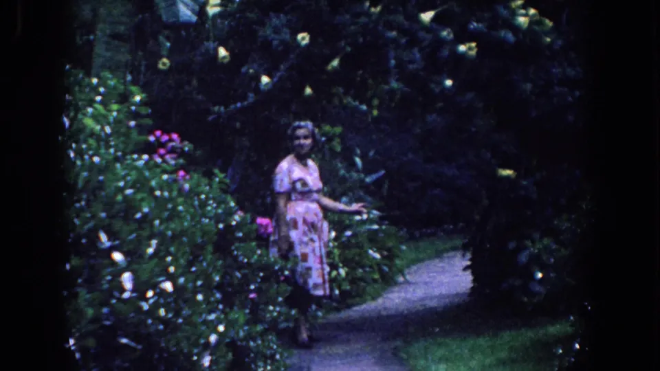 The 1958 Florida footage shows the woman walking along a curved garden path with flowers and heavy foliage nearby.
