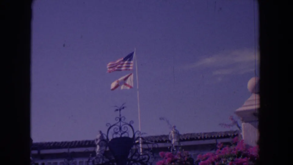 1958 Florida frame showing American and Florida state flags on a flagpole above an ornate iron gate, with flowers at the lower edge.