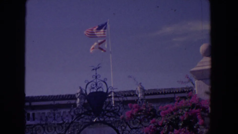 1958 Florida home-movie frame with two flags raised over decorative ironwork, a roofline, blue sky, and pink bougainvillea-like flowers.