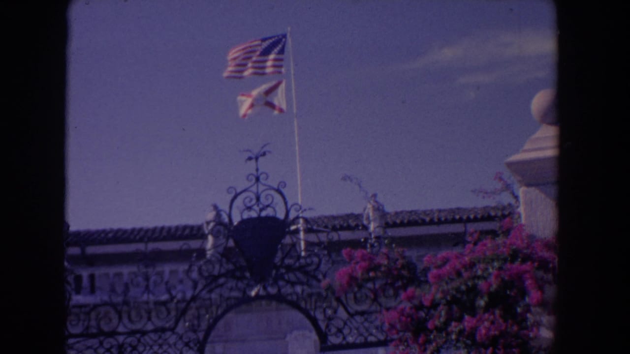 1958 Florida home-movie frame showing two flags above decorative ironwork, a tiled roofline, pale arches, and bougainvillea.