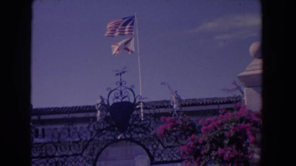 1958 Florida home-movie frame showing two flags above decorative ironwork, a tiled roofline, pale arches, and bougainvillea.