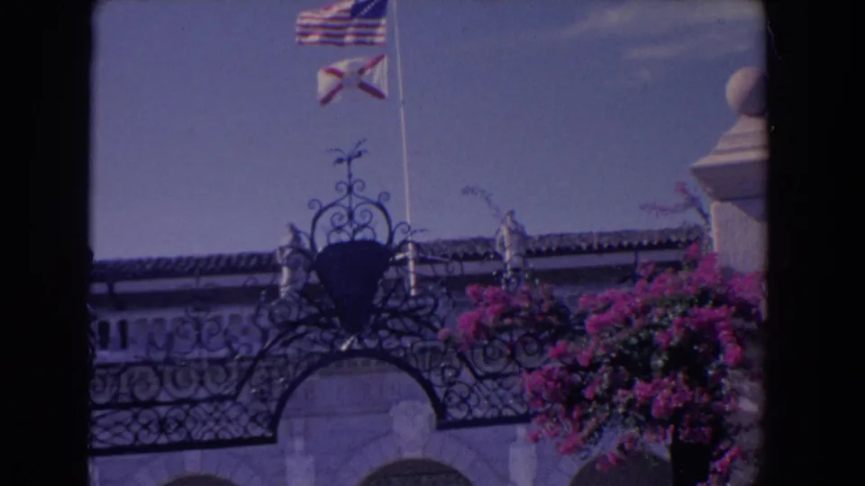 1958 Florida exterior frame with flags above an ornate roofline, pale architectural details, and bright pink bougainvillea at right.