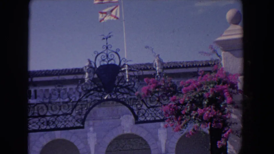 1958 Florida home-movie view of ornate ironwork over arched masonry with a flagpole, tiled roof, and bougainvillea blooms.