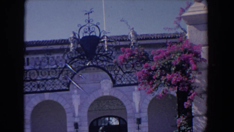 1958 Florida frame showing ornate iron roof detail over pale arched architecture, with bougainvillea and a partial flagpole nearby.