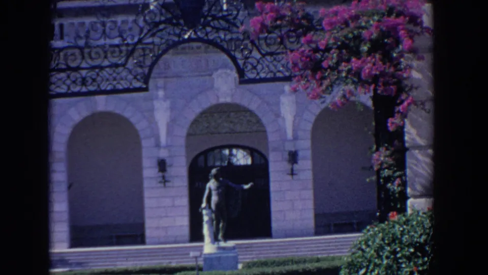 1958 Florida 8mm frame showing the entrance steps, hedge, statue, arched openings, and bougainvillea on the facade.