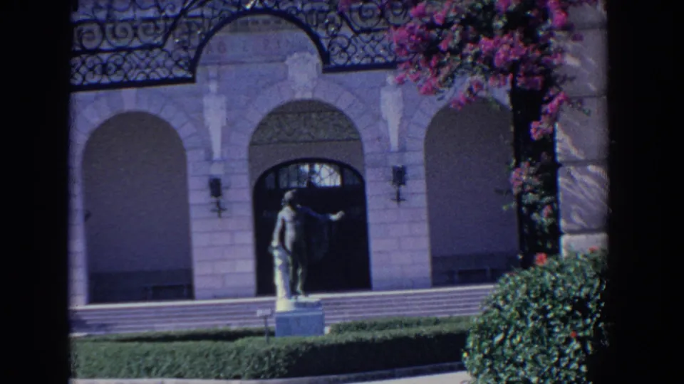 1958 Florida home-movie frame of the ornate entrance, showing statue, steps, hedge, arched facade, and vivid bougainvillea.