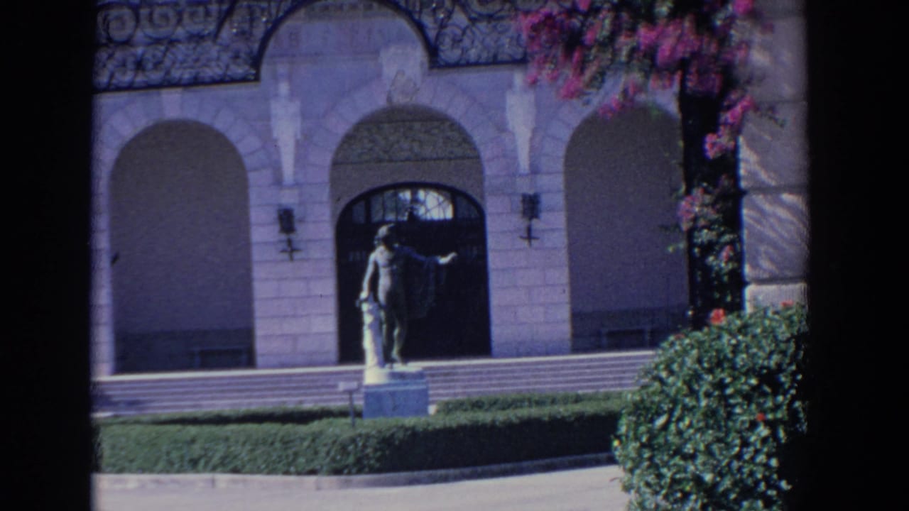 1958 Florida 8mm frame showing a mansion facade with arched entryways, a classical statue, trimmed lawn, and flowering branches.