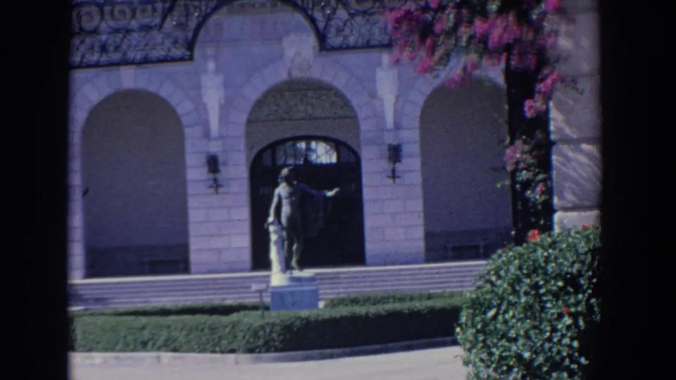 1958 Florida 8mm frame showing a mansion facade with arched entryways, a classical statue, trimmed lawn, and flowering branches.