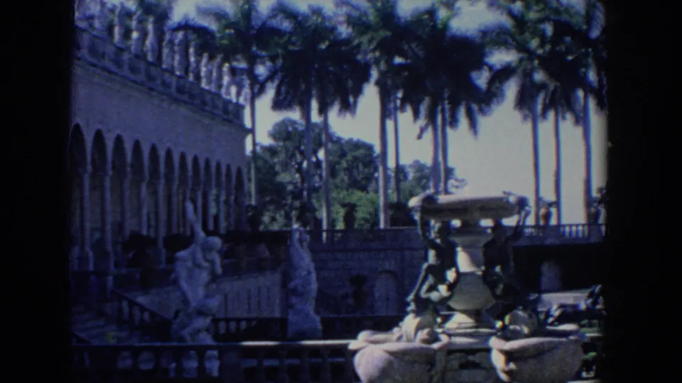 1958 Florida 8mm frame looking across mansion grounds with tall palms, terrace railings, sculptures, and an ornate fountain basin.