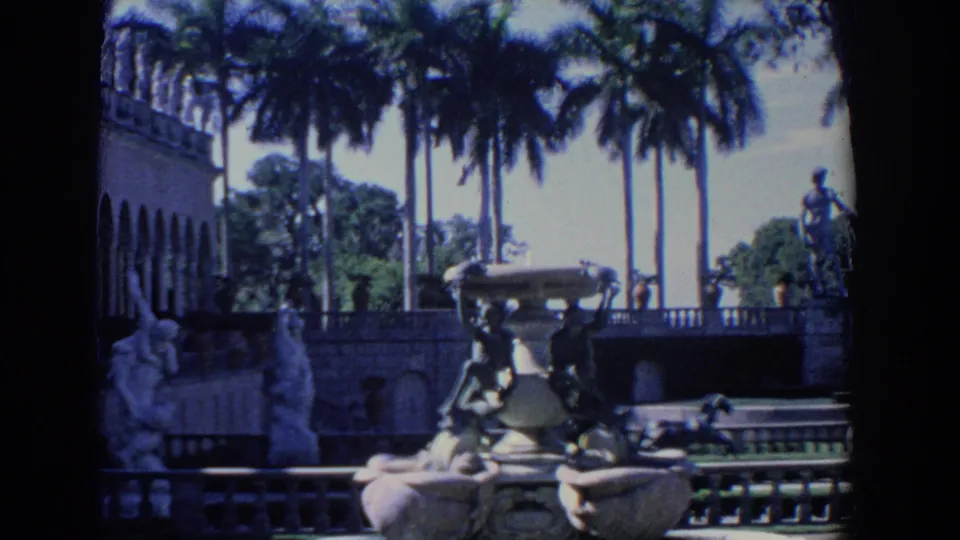 1958 Florida 8mm frame of estate grounds with palms, a large decorative fountain, sculpture figures, railings, and arched architecture.