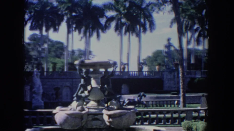 1958 Florida 8mm frame centered on an ornate estate fountain, with palms, a stone railing, lawn, shrubs, and a standing statue.