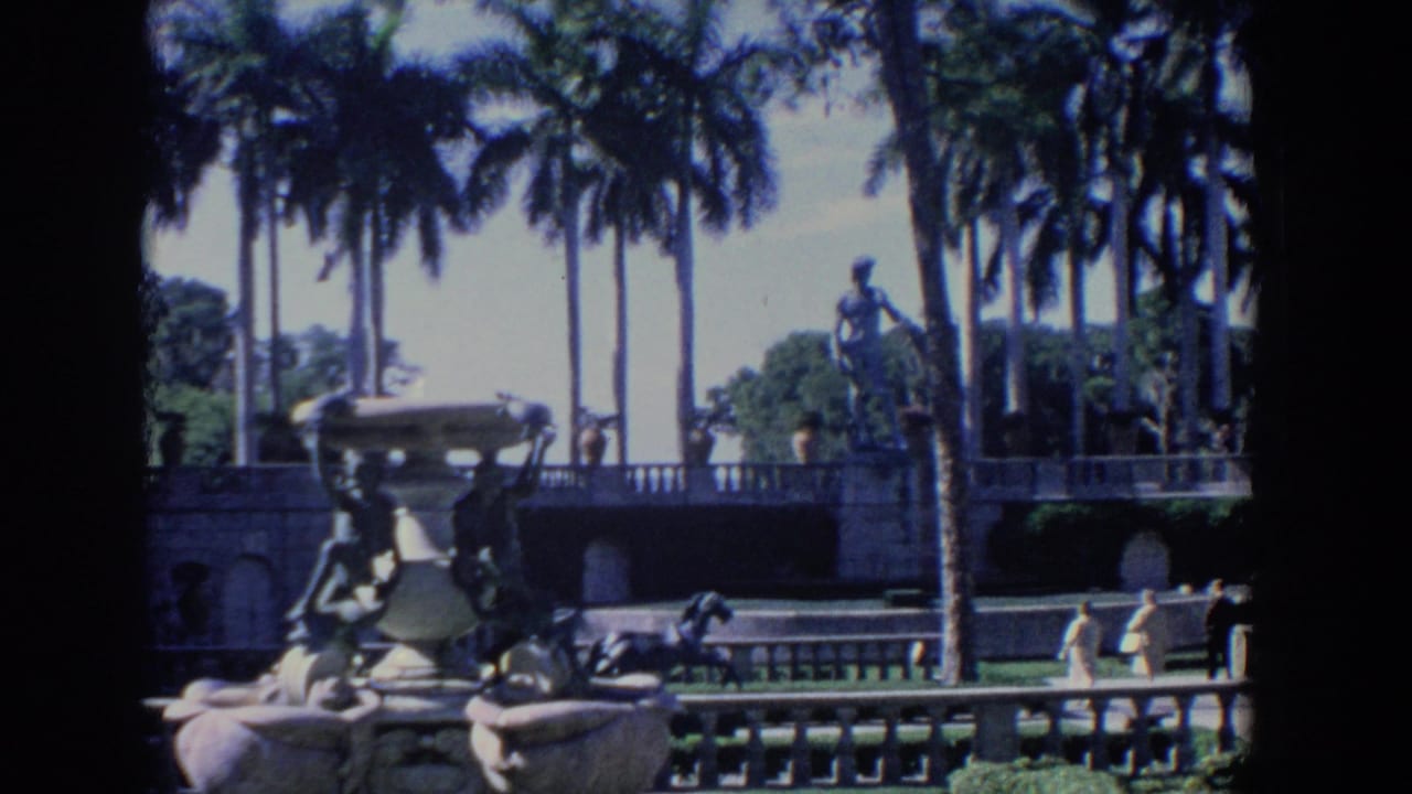 1958 Florida 8mm frame showing visitors walking through a formal palm-lined garden with stone fountain figures and sculpture.