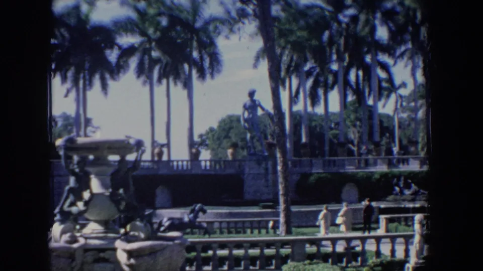 1958 Florida garden view with a stone fountain at left, tall palms, a raised statue, and distant visitors on the walkway.