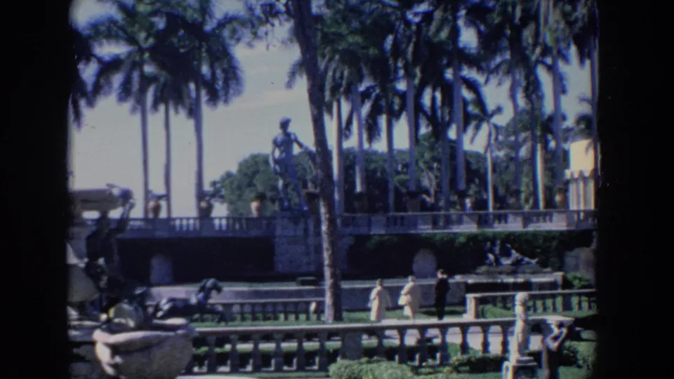 1958 Florida 8mm frame with visitors in light clothing walking along a formal garden terrace beneath tall palms and statuary.