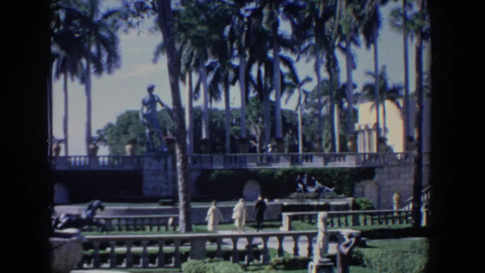 1958 Florida travel footage frame showing distant visitors on a terraced walkway, with palm trees, stone railings, and sculpture behind them.