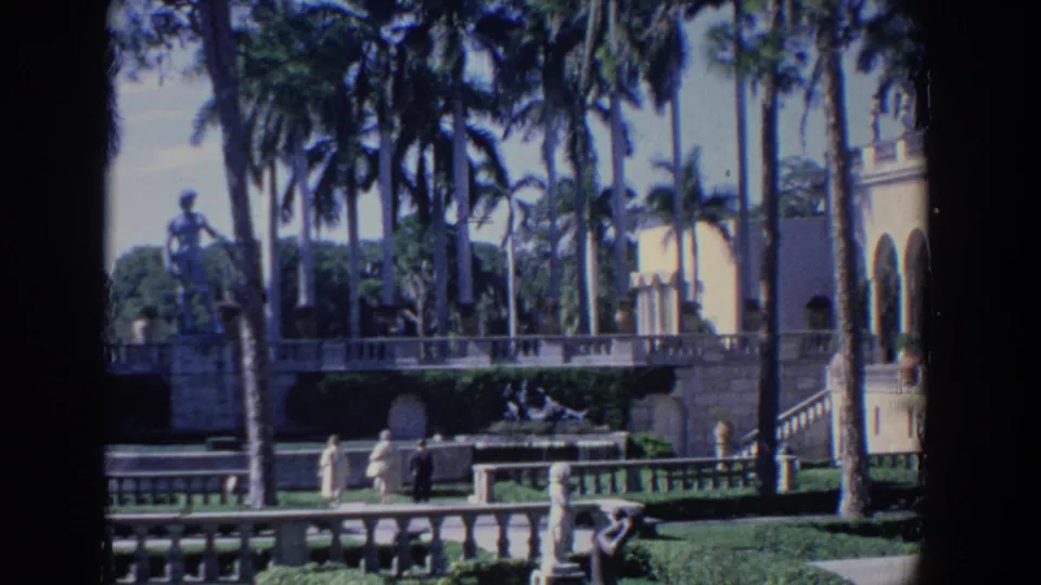 Florida estate garden view with palms, a white arched building, stone railings, statues, and distant visitors.