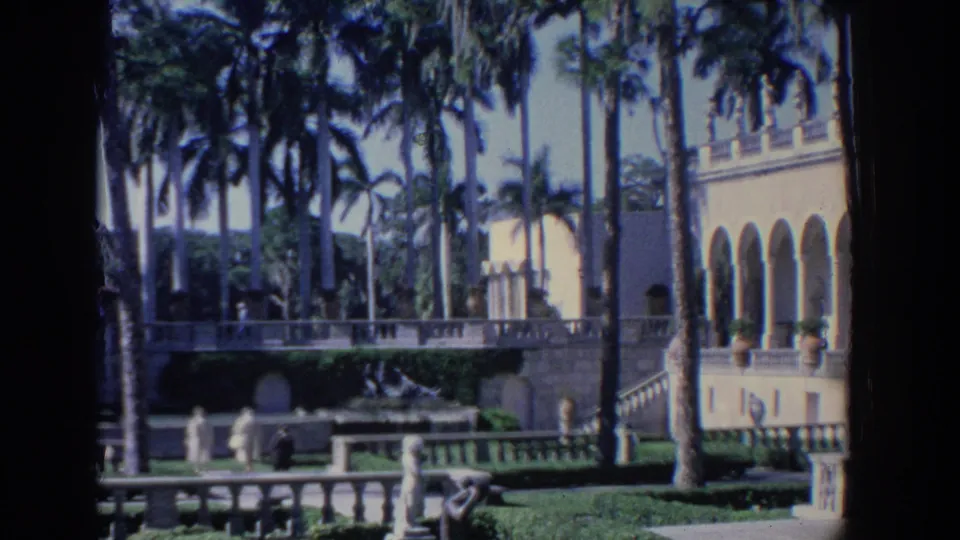 1958 Florida home-movie frame showing palms, formal terraces, statues, and a white building with arched colonnades.