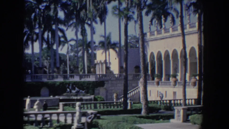 Florida 1958 8mm still of a white arched estate facade beside palms, balustrades, statues, and strolling visitors.