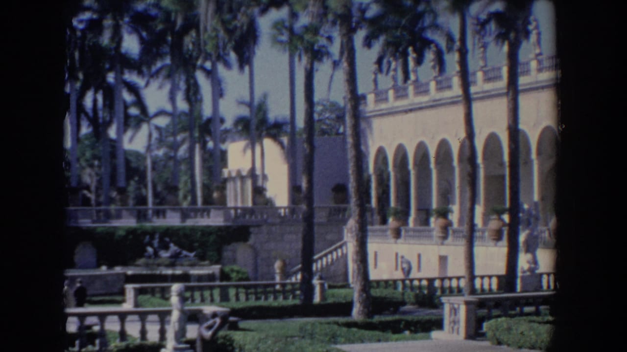 1958 Florida 8mm frame showing a grand estate facade with arched colonnades, palms, stone railings, and manicured grounds.