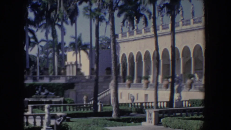 1958 Florida home-movie view of a large mansion facade with palms, arched walkway, balustrades, and clipped garden areas.