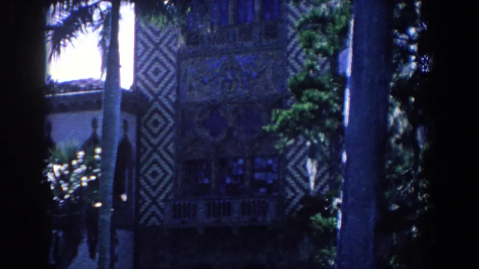 1958 Florida home-movie frame with tall palms, deep shadows, and an ornate building facade with geometric wall decoration.