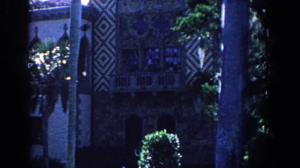 1958 Florida frame showing palms and shrubbery before an ornate building with windows, balcony railing, and diamond-patterned exterior details.
