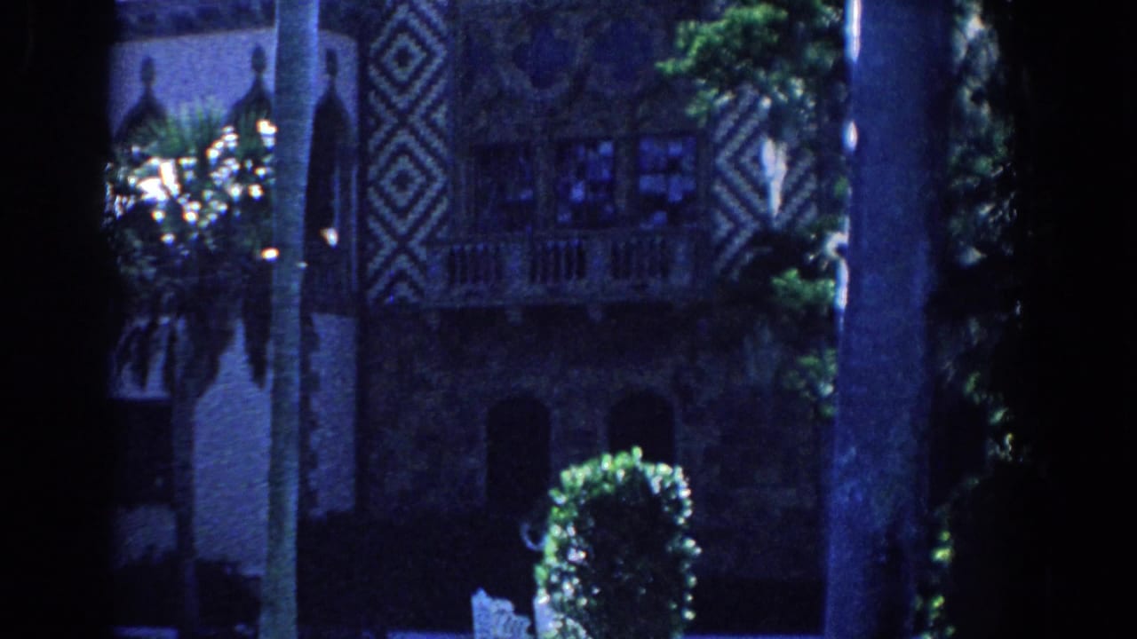 1958 Florida 8mm frame showing a dark estate exterior framed by palms, geometric facade panels, lawn, hedge, and garden chair.