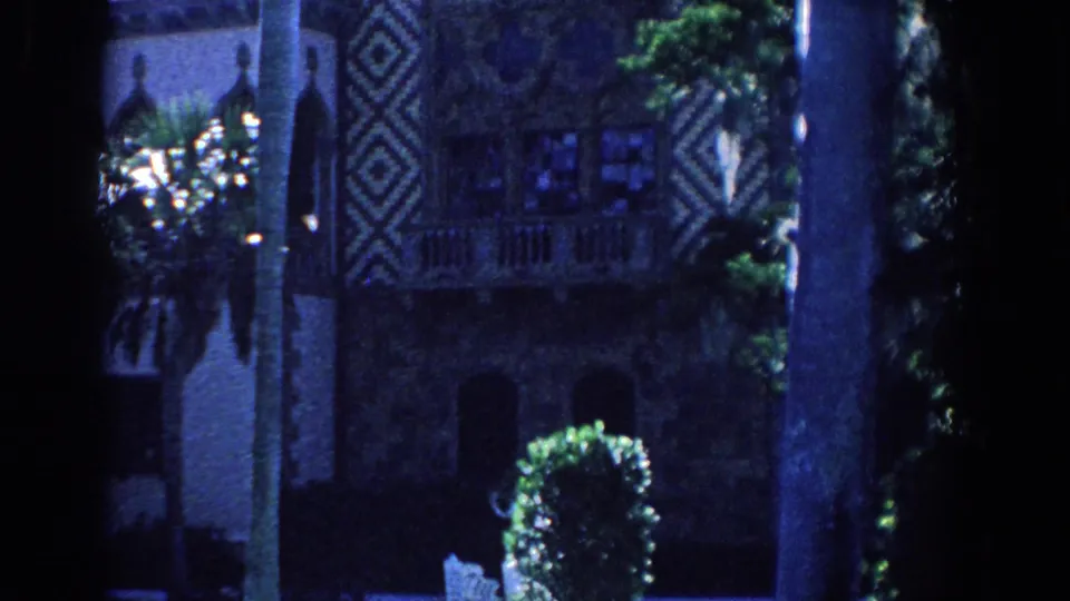 1958 Florida 8mm frame showing a dark estate exterior framed by palms, geometric facade panels, lawn, hedge, and garden chair.