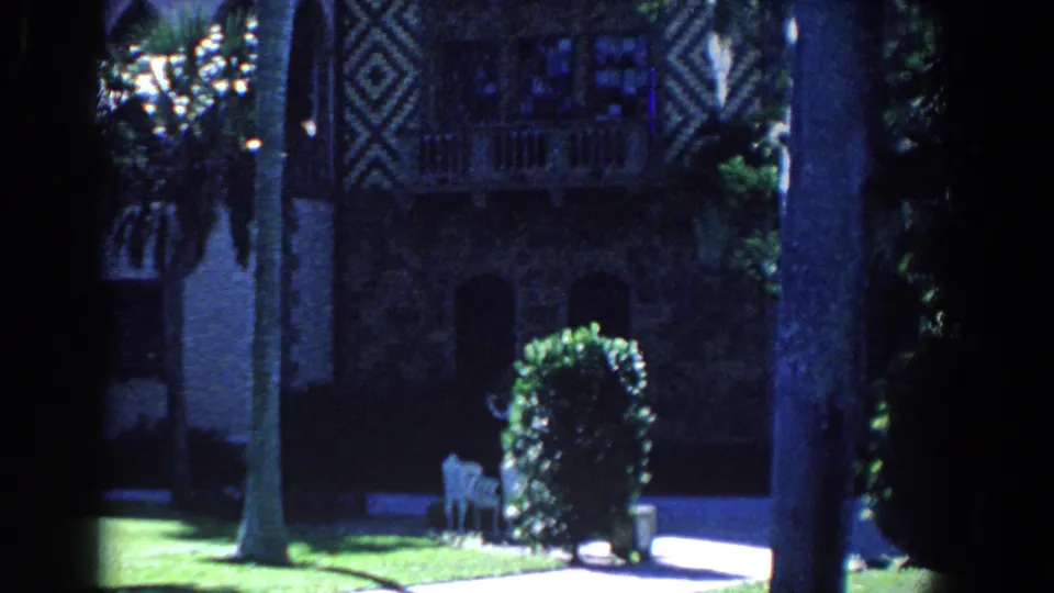 1958 Florida home-movie frame with a shaded estate wall, balcony-like detail, palm trunks, lawn, hedge, and white garden chair.