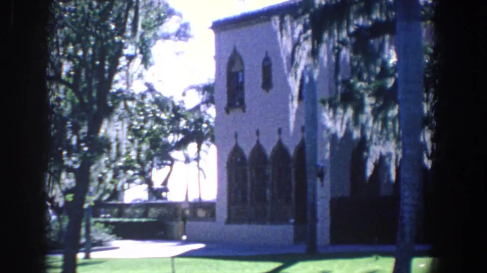 1958 Florida 8mm frame showing the side of a pale arched building beside palms, trees, walkway, and bright outdoor light.