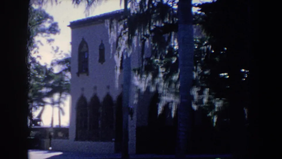 1958 Florida home-movie frame of a shaded arched building side, with palm trunks and trees silhouetted against bright exterior light.
