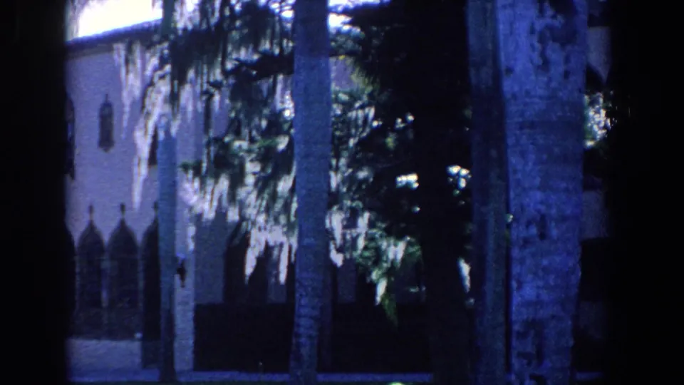 1958 Florida travel footage frame with a palm trunk, dense foliage, and a partially visible Gothic-style stucco facade.