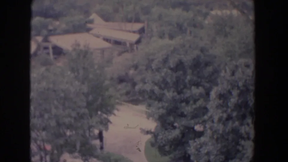 1958 Florida 8mm frame showing brown rooftops, leafy grounds, a pale path, and a tiny distant person near the lower edge.