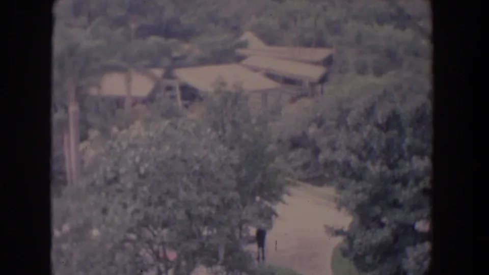 1958 Florida 8mm frame with low brown rooftops, heavy tree cover, and a distant person walking on a pale curved path.