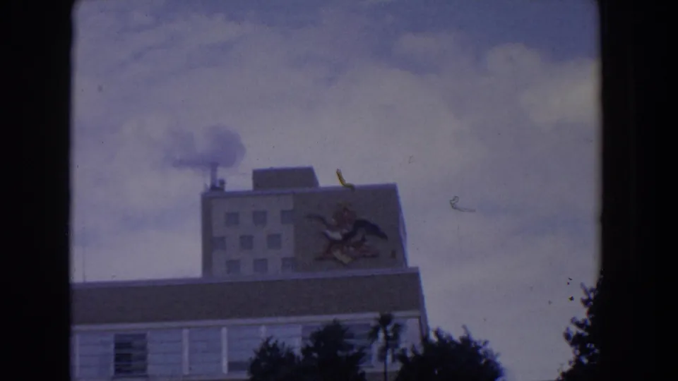 1958 Florida 8mm frame showing a large building, smoke from a rooftop chimney, and a decorative bird mural on the facade.